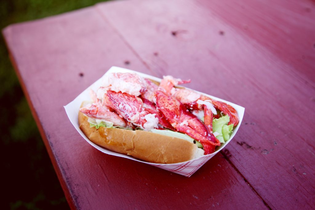 A delectable lobster roll filled with succulent lobster meat, nestled on a crisp, golden bun, served on a rustic picnic table under a bright, sunny day.